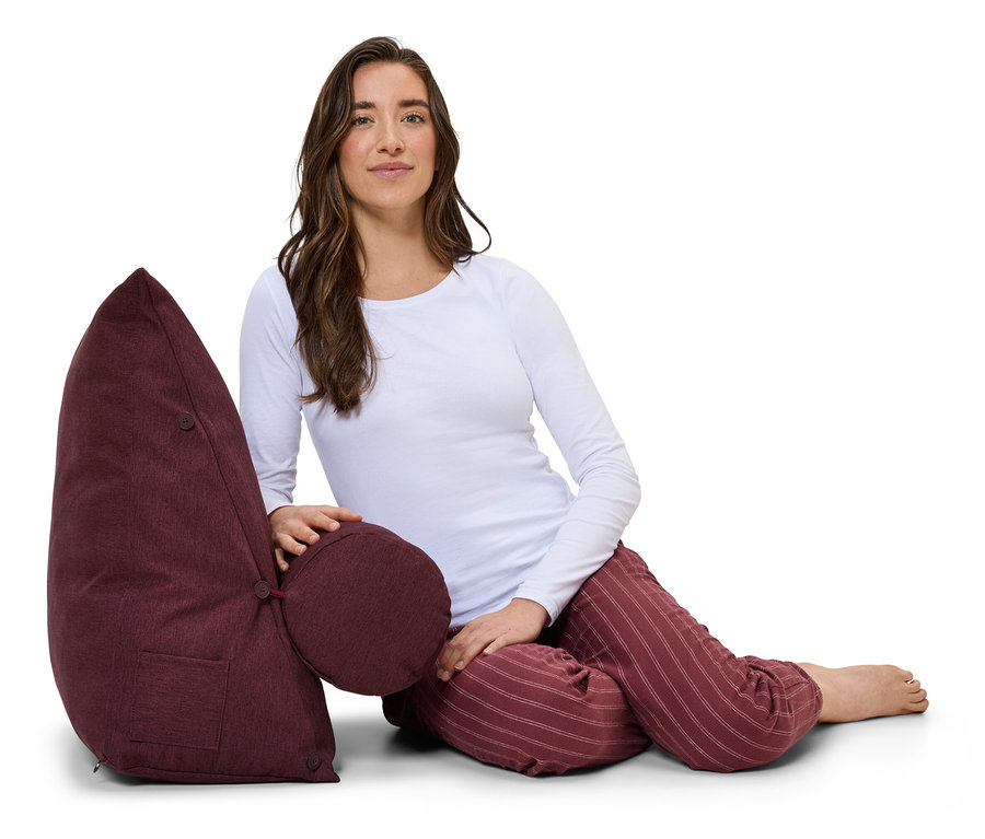 Frau sitzt neben weinrotem Entspannungskissen mit Rücken- und Lendenwirbelstütze.