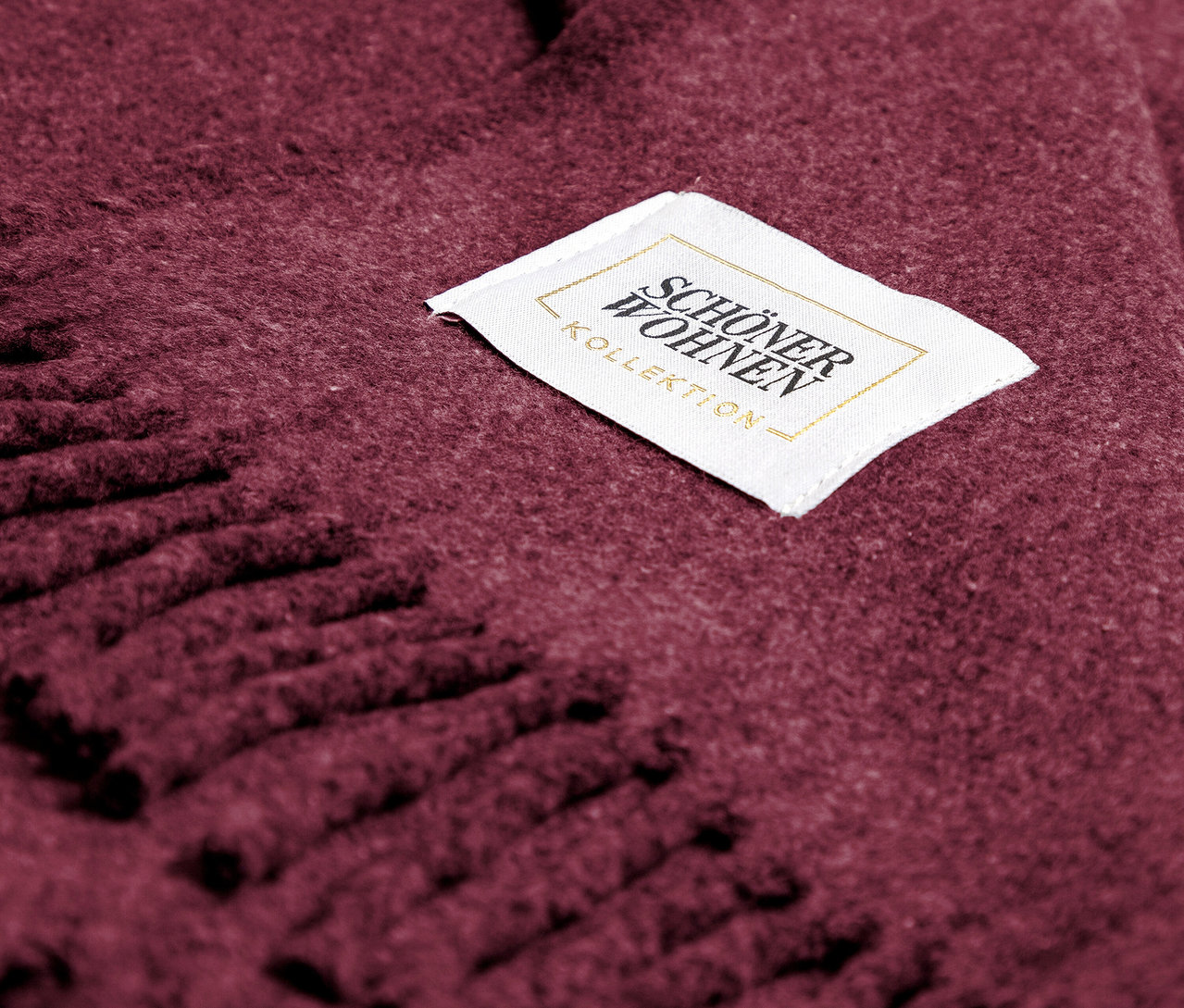 Nahaufnahme einer bordeauxroten Decke mit Fransen und einem weißen Etikett mit der Aufschrift SCHÖNER WOHNEN - KOLLEKTION.