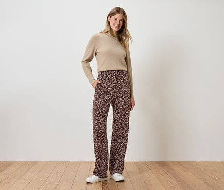 Frau steht mit Händen in den Hosentaschen, trägt eine braune, geblümte Hose und einen beigen Pullover.