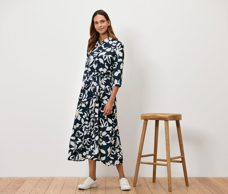 Eine Frau steht neben einem Holzhocker und trägt ein marineblaues Kleid mit weißem Blumenmuster.