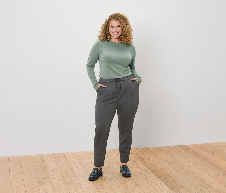 Frau mit lockigen blonden Haaren, die ein grünes Langarmshirt und eine graue Hose mit ihren Händen in den Taschen trägt.