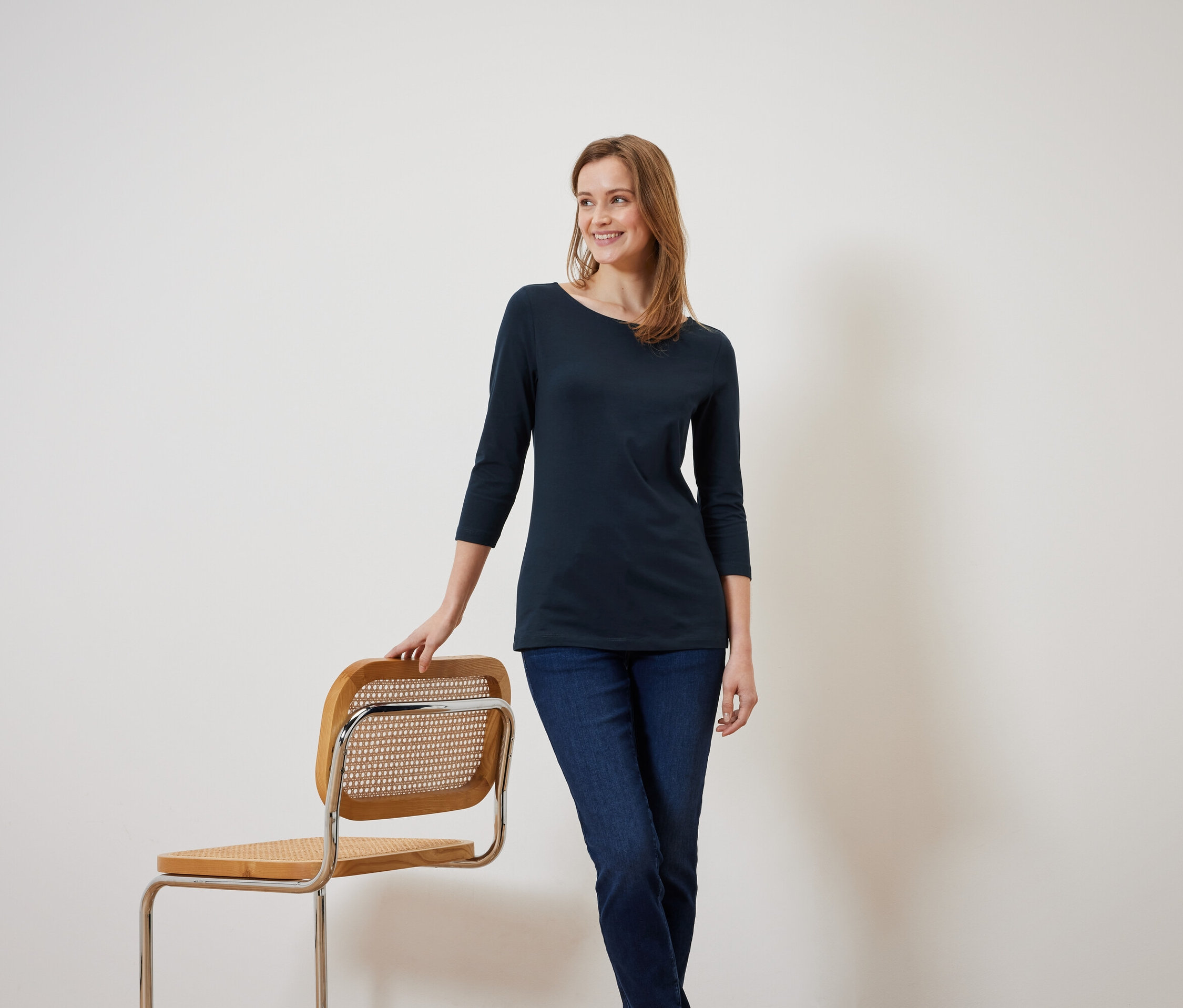 Eine lächelnde Frau in einer marineblauen Bluse mit Dreiviertelärmeln lehnt an einem Holzstuhl.