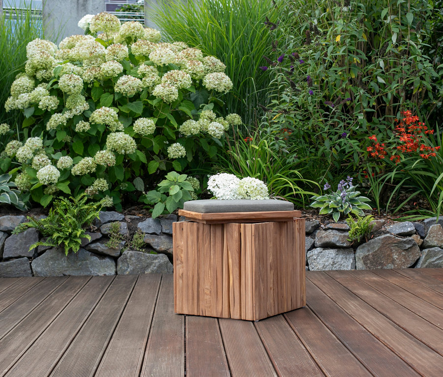 Ein deVries Sitzhocker mit Stauraum steht auf einer Holzterrasse vor einer Steinmauer und Pflanzen.