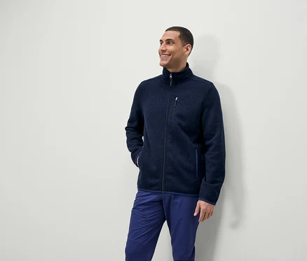 Ein Mann lächelt und schaut zur Seite, er trägt eine blaue Fleecejacke mit Reißverschluss und blaue Hosen.