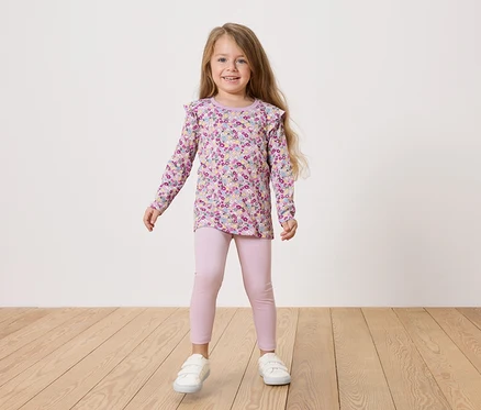 Ein Mädchen mit blonden Haaren trägt ein geblümtes Hemd und rosa Leggings und weiße Schuhe.