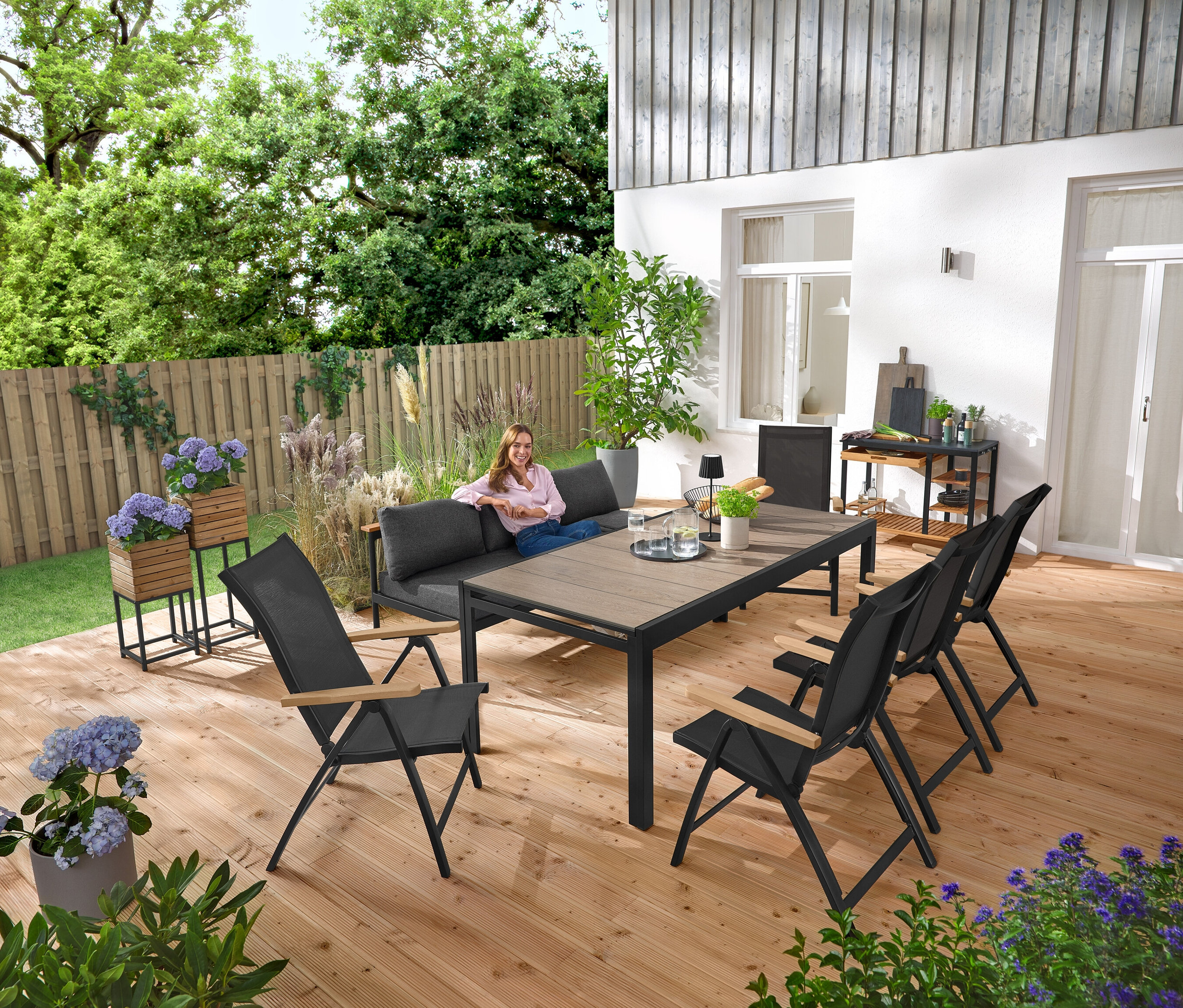 Eine Frau sitzt auf einer 2-in-1-Gartenbank ‒ Sonnenliege »Elin«. Im Hintergrund stehen 2 Pflanzsäulen »Elin«. Auf der Terrasse stehen auch ein Grillwagen mit Polyzement-Arbeitsfläche und ein ausziehbarer Gartentisch mit Duraboard-Tischplatte.