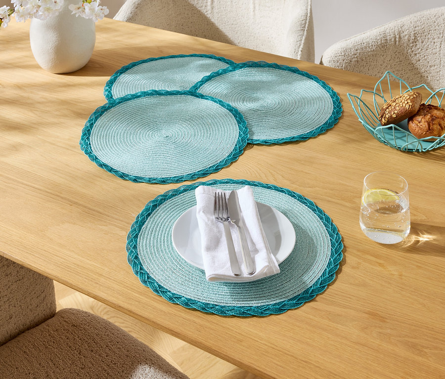 Ein Esstisch mit vier türkisfarbenen Tischsets, einem Teller mit Besteck, einem Glas Wasser, einer Blumenvase und einem Korb mit Brot.