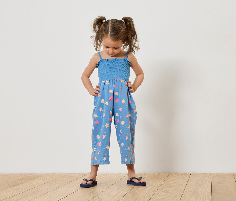Mädchen mit Zöpfen steht mit Händen in den Hüften, trägt einen blauen Jumpsuit mit Muschel- und Seesternmuster und blaue Flip-Flops.