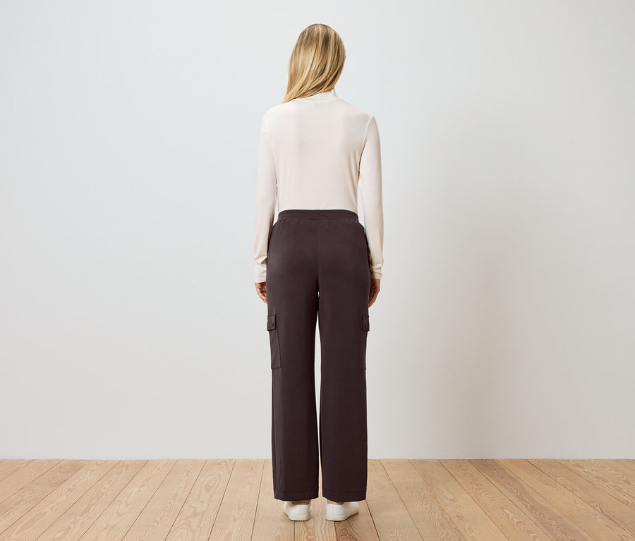 Rückansicht einer Frau mit blonden Haaren, die einen cremefarbenen Rollkragenpullover, braune Cargohose und weiße Turnschuhe trägt.