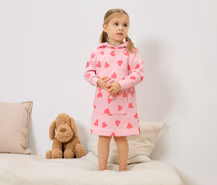 Ein kleines Mädchen mit blonden Zöpfen steht auf einem Bett und trägt ein rosafarbenes Kapuzenkleid mit roten Herzen. Neben ihr sitzt ein Stoffhund.