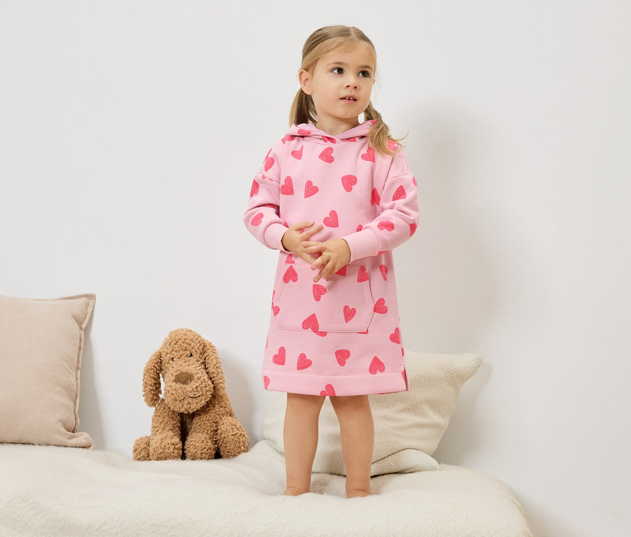 Ein kleines Mädchen mit blonden Zöpfen steht auf einem Bett und trägt ein rosafarbenes Kapuzenkleid mit roten Herzen. Neben ihr sitzt ein Stoffhund.