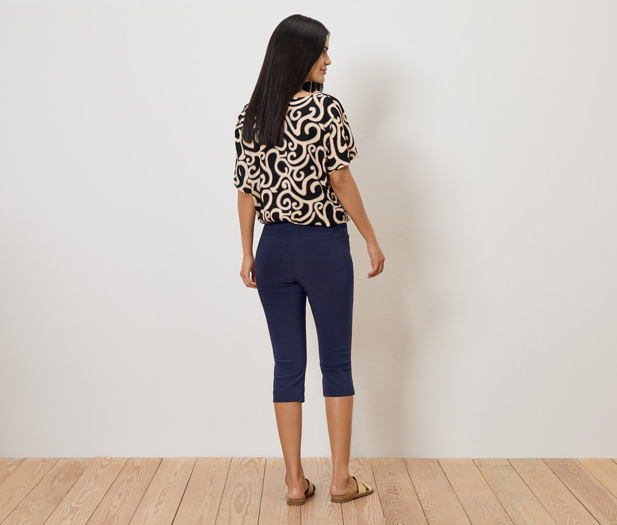 Frau von hinten in gemustertem Oberteil, blauen Caprihosen und Sandalen vor einer weißen Wand.