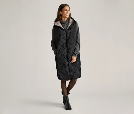 Eine Frau steht und lächelt, sie trägt eine lange, schwarze Steppweste mit Kapuze über einem grauen Pullover, Strumpfhosen und Stiefeln.