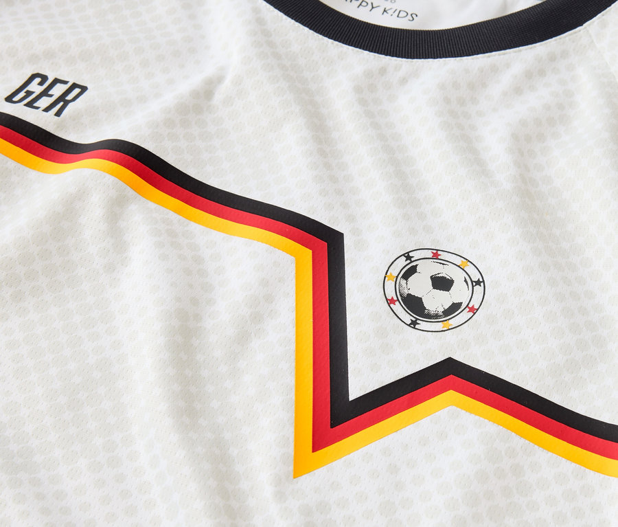 Detailansicht eines weißen Trikots mit dem Aufdruck "GER", einem Fußball-Patch und schwarz-rot-goldenen Streifen.