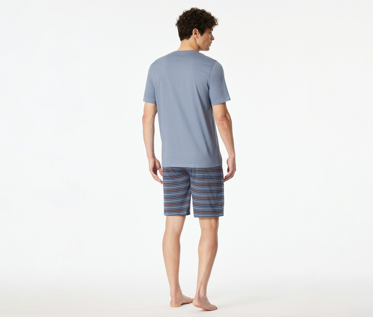 Ein Mann von hinten, der ein hellblaues T-Shirt und gestreifte Shorts des SCHIESSER Shorty-Sets trägt. Er ist barfuß.