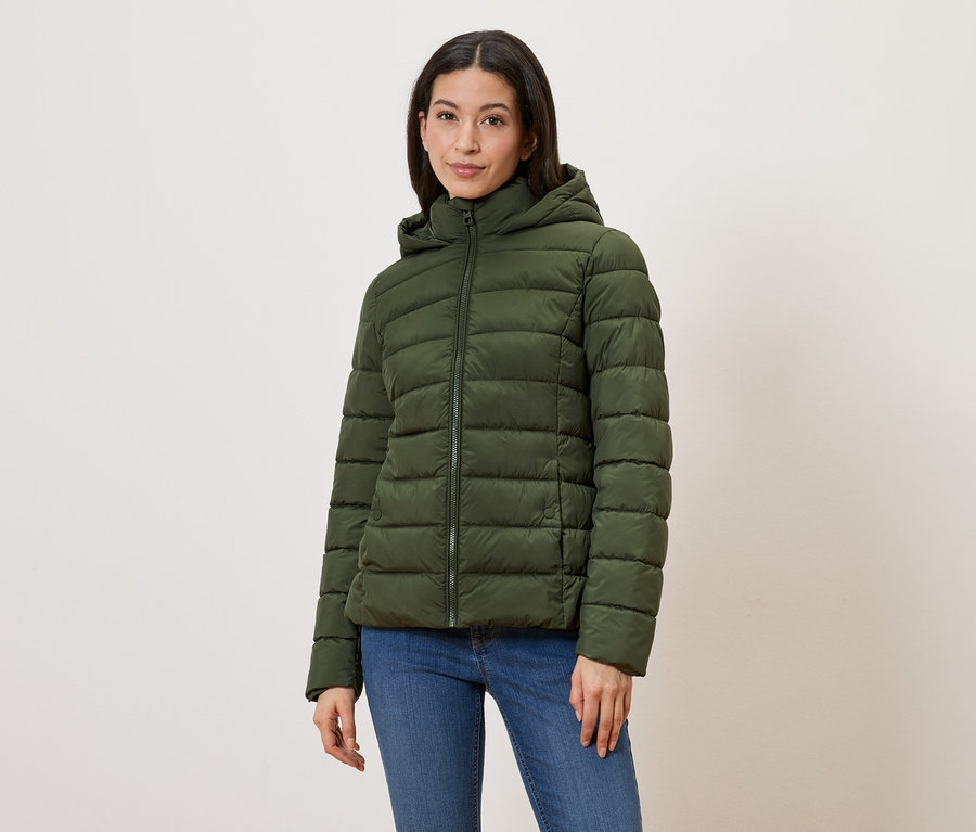 Frau mit olivgrüner Steppjacke mit Kapuze und Reißverschluss, dazu blaue Jeans vor hellem Hintergrund.