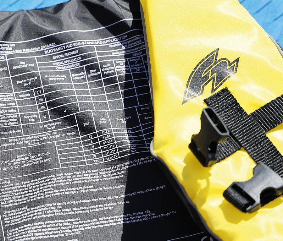 Eine gelbe und graue Schwimmweste mit schwarzen Gurten und Schnallen liegt neben einem schwarzen Etikett mit Warnhinweisen und einer Größentabelle.