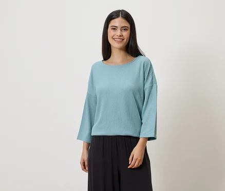 Frau mit langen, dunklen Haaren, die ein hellblaues, langärmeliges Oberteil und eine schwarze Hose vor einem weißen Hintergrund trägt.