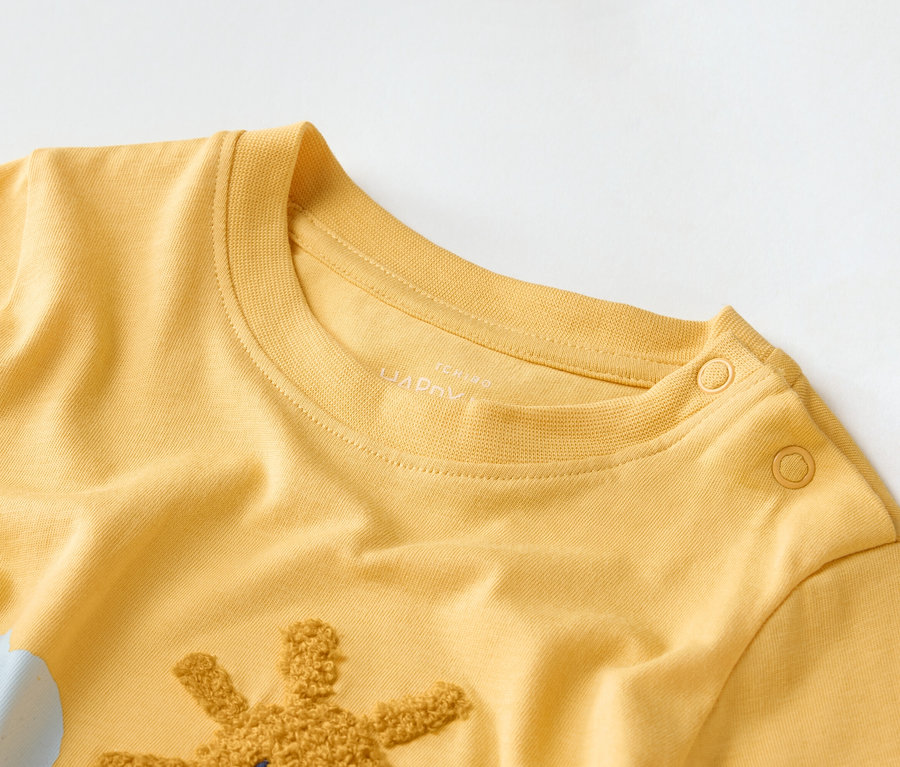 Detailaufnahme eines goldgelben Kinder-T-Shirts mit Giraffen-Applikation und Wolkenmotiv.