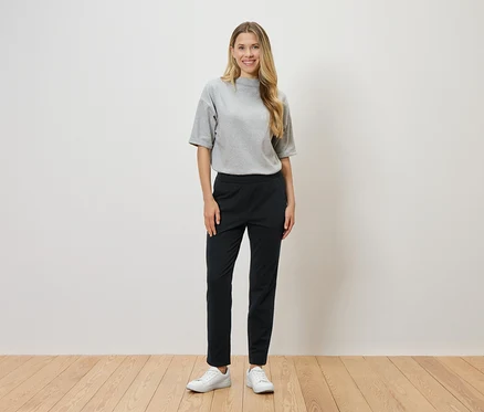 Eine Frau mit blonden Haaren steht vor einer weißen Wand und trägt ein graues T-Shirt, eine schwarze Hose und weiße Turnschuhe.