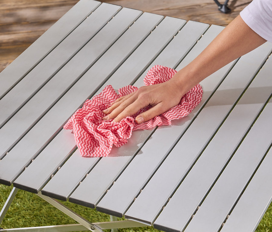 Eine Hand wischt mit einem rot-weiß karierten Tuch über einen grauen Campingtisch.
