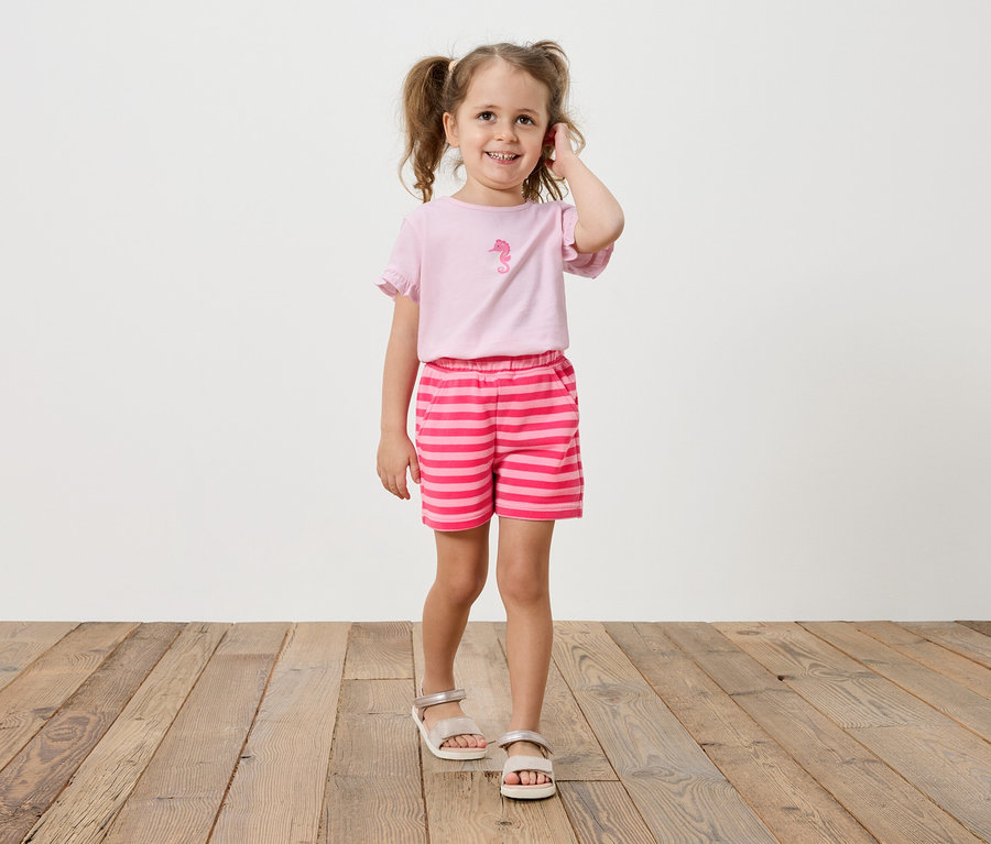 Mädchen mit Zöpfen trägt ein rosa T-Shirt mit einem Seepferdchen-Aufdruck und gestreifte Shorts.