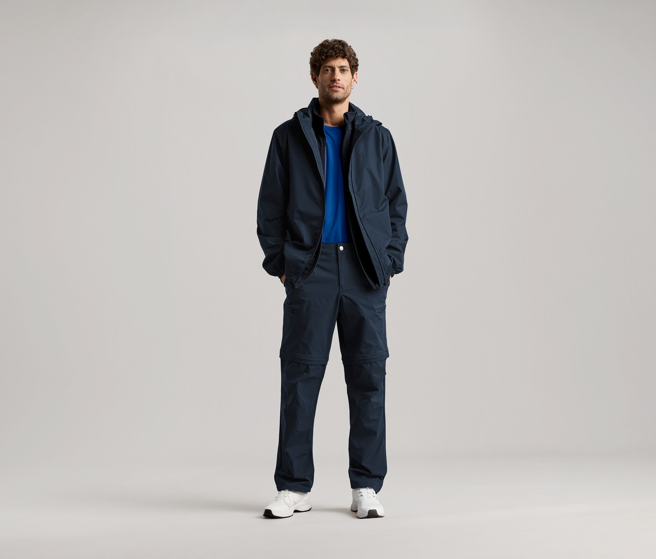 Ein Mann steht mit seinen Händen in den Taschen einer dunkelblauen Jacke und einer passenden Hose, dazu trägt er ein blaues T-Shirt und weiße Turnschuhe.