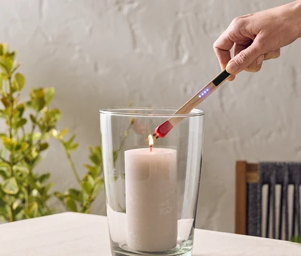 Eine Hand zündet eine Kerze in einem Glas mit einem Stabfeuerzeug an. Das Glas steht auf einem Tisch.