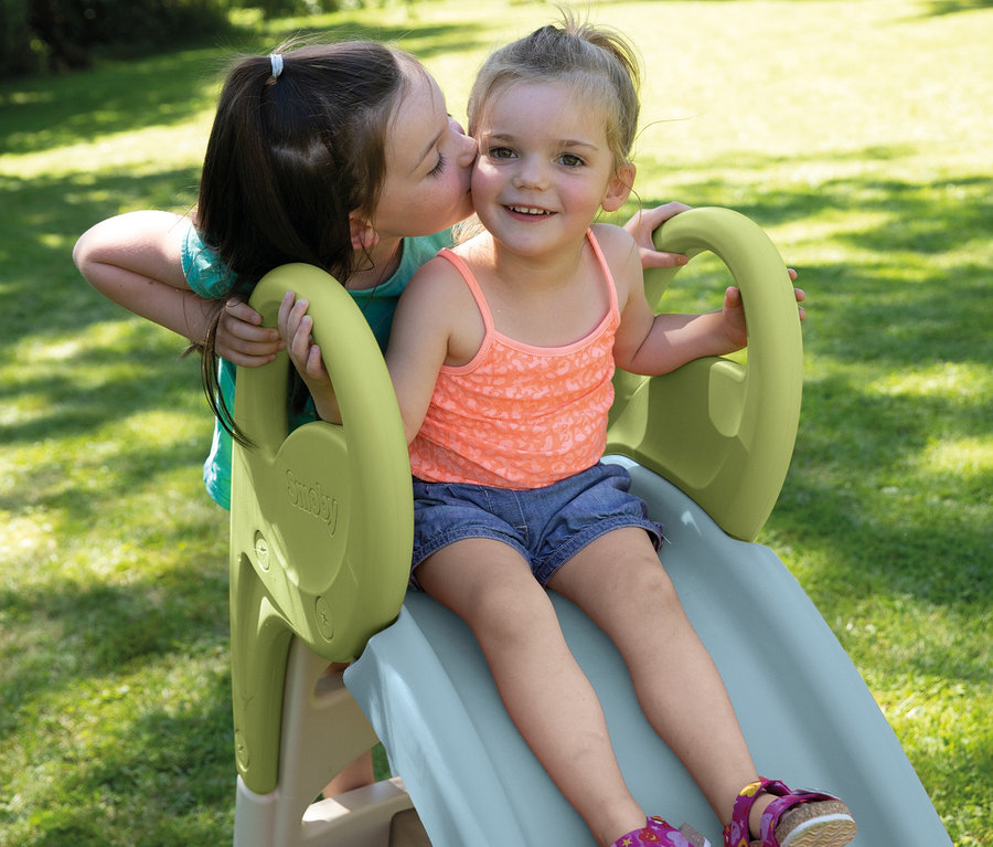 Mädchen gibt einem anderen Mädchen, das auf der Smoby Life Rutsche »GM« sitzt, einen Kuss auf die Wange.
