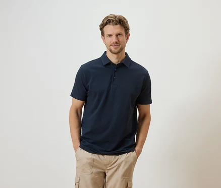 Mann mit blonden Haaren und Bart, bekleidet mit einem dunkelblauen Poloshirt und beigen Cargohosen, posiert vor einem weißen Hintergrund.