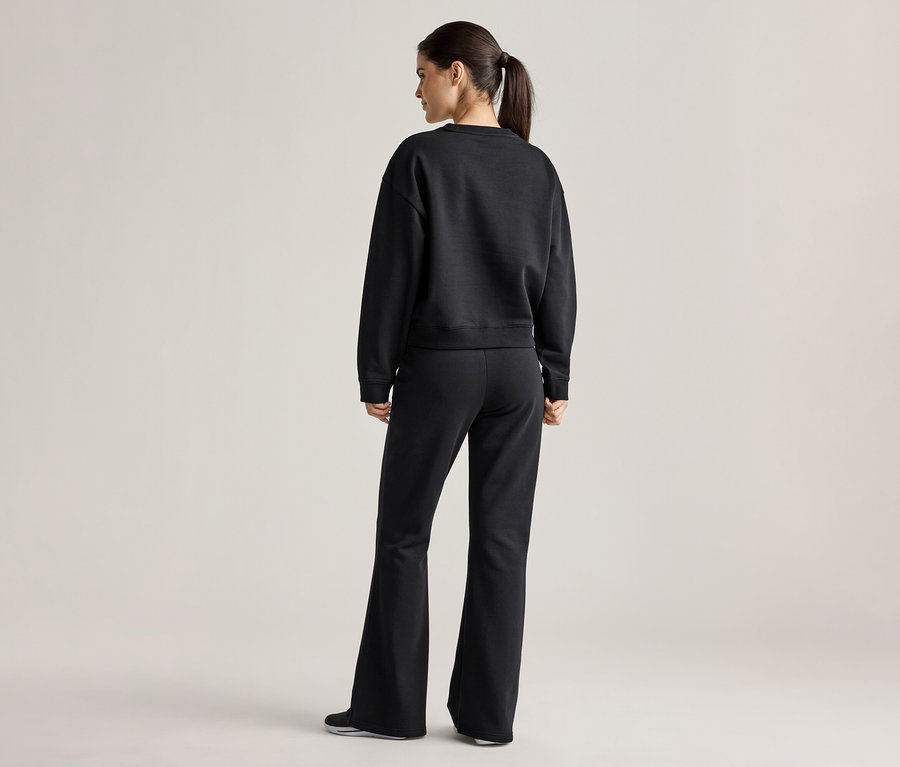 Rückansicht einer Frau mit Pferdeschwanz, die einen schwarzen Pullover und eine ausgestellte Hose trägt.