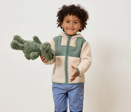 Ein lächelnder Junge mit lockigem Haar hält ein grünes Stofftier-Dinosaurier und trägt eine hellbraune Jacke mit grünen Akzenten und blaue Hosen.