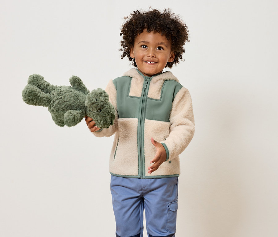 Ein lächelnder Junge mit lockigem Haar hält ein grünes Stofftier-Dinosaurier und trägt eine hellbraune Jacke mit grünen Akzenten und blaue Hosen.