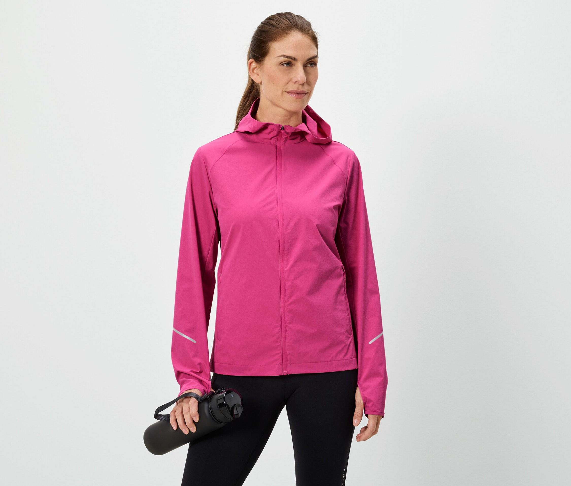Frau hält eine schwarze Wasserflasche, trägt eine pinke Jacke und schwarze Leggings vor einem weißen Hintergrund.