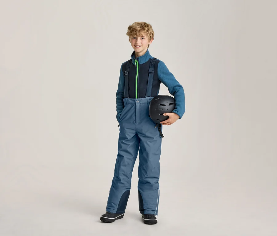 Lächelnder Junge in blauer Skihose mit schwarzen Hosenträgern und einer blau-schwarzen Fleecejacke, der einen schwarzen Skihelm hält.
