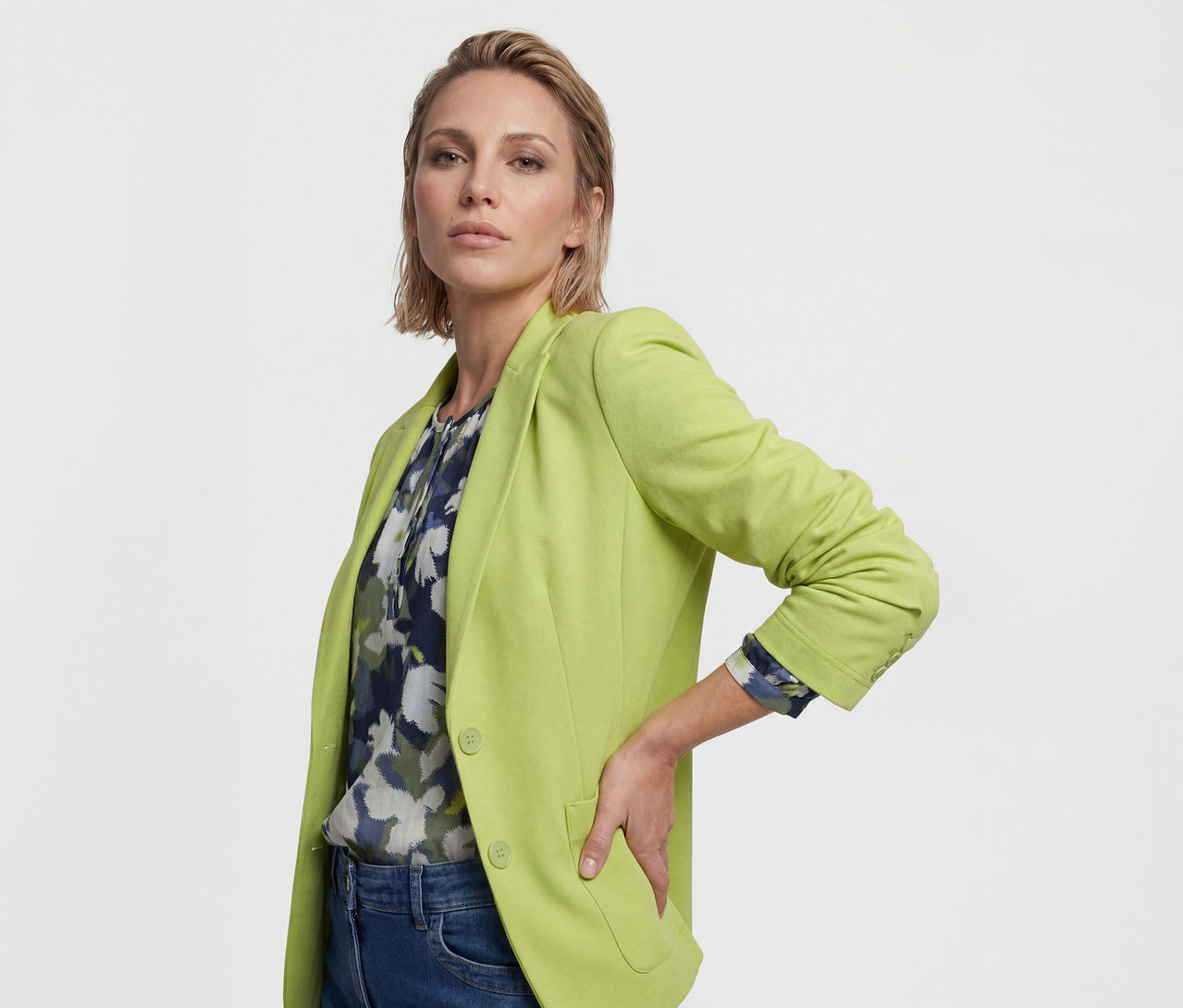 Eine Frau mit kurzen, hellbraunen Haaren trägt einen limettengrünen Blazer über einer dunkelblauen Blumenbluse und blauen Jeans, die Hand in die Hüfte gestützt.