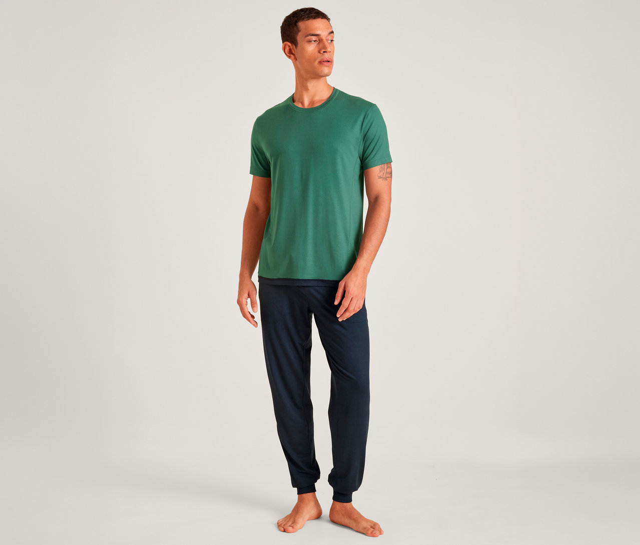 Mann trägt ein grünes T-Shirt und eine dunkelblaue Jogginghose, steht barfuß und blickt nach rechts.