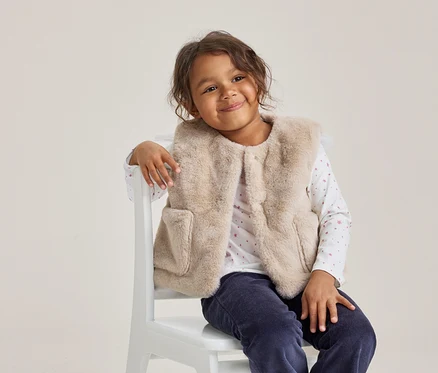 Junges Mädchen mit dunklen Locken lächelt, trägt eine hellbeige Kunstfellweste, ein weißes Langarmshirt mit rosa Mustern und eine dunkelblaue Hose, sitzt auf einem weißen Stuhl.