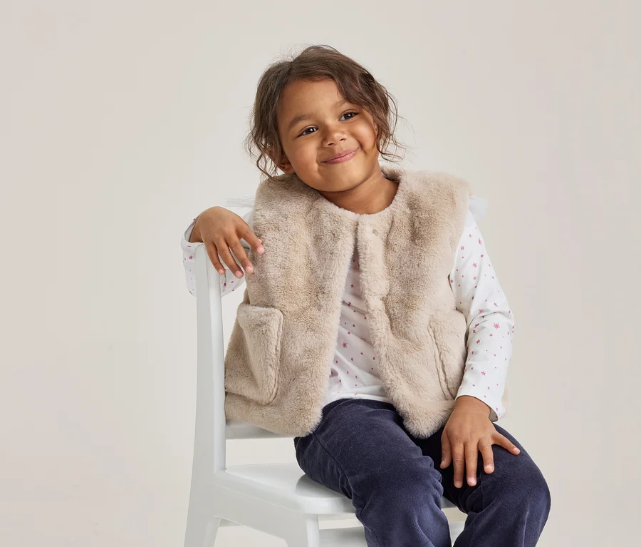 Junges Mädchen mit dunklen Locken lächelt, trägt eine hellbeige Kunstfellweste, ein weißes Langarmshirt mit rosa Mustern und eine dunkelblaue Hose, sitzt auf einem weißen Stuhl.