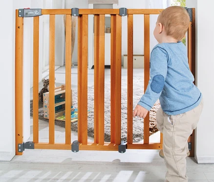 Kleinkind in blauem Langarmshirt und beiger Hose blickt durch ein roba Türschutzgitter »Safety Up« aus Holz.