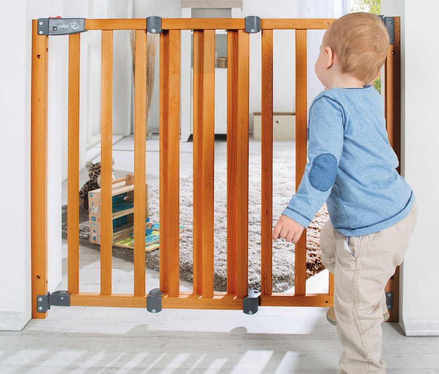 Kleinkind in blauem Langarmshirt und beiger Hose blickt durch ein roba Türschutzgitter »Safety Up« aus Holz.