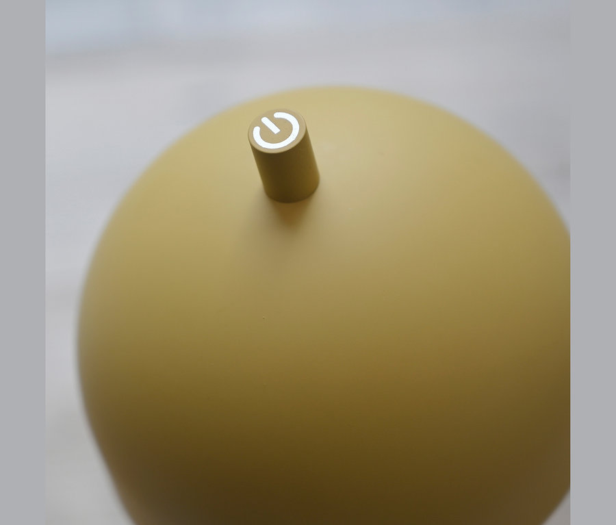 Nahaufnahme eines matten, gelben, kugelförmigen Objekts mit einem kleinen zylindrischen Knopf oben, der ein weißes Einschaltsymbol zeigt.