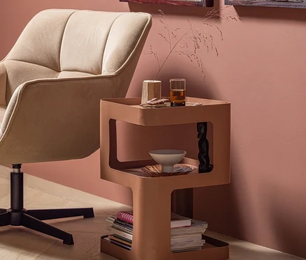 Beiger Polstersessel neben einem braunen, mehrstufigen Beistelltisch. Auf dem Tisch stehen ein Glas mit Getränk, Dekoartikel und Zeitschriften. Der Raum hat eine rosa Wand und einen hellen Holzfußboden.