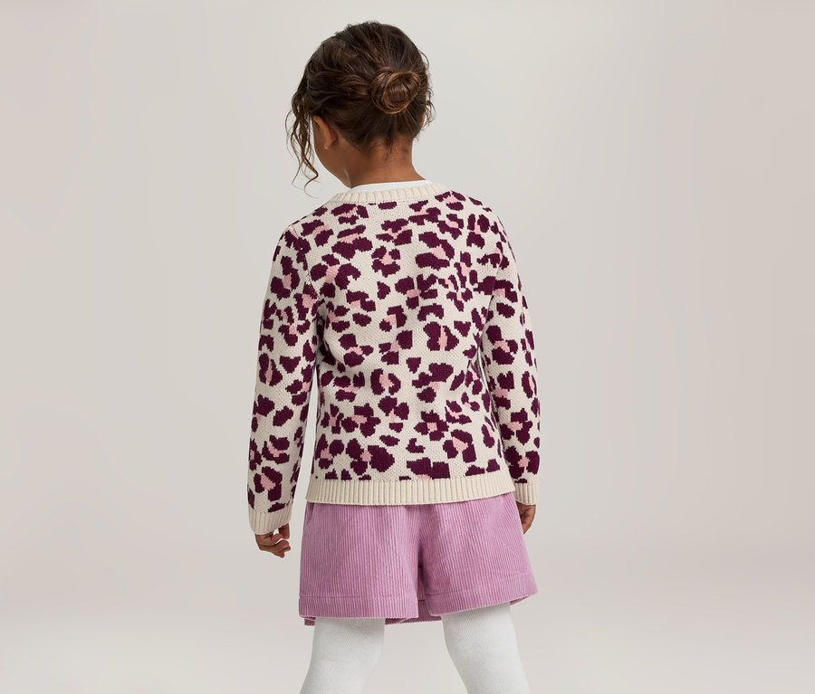 Ein junges Mädchen von hinten gesehen, das einen cremefarbenen und lila Leopardenmuster-Cardigan, lila Cord-Shorts und weiße Strumpfhosen trägt. Ihr dunkles Haar ist zu einem Dutt gebunden.