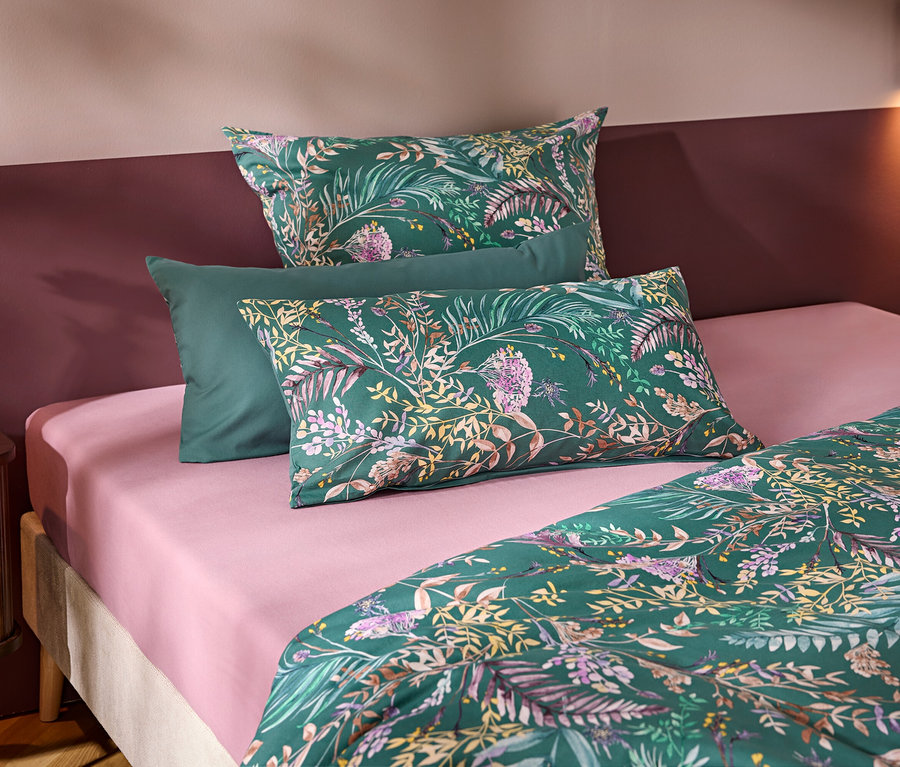 Ein Bett mit einem rosa Spannbettlaken und grüner Bettwäsche mit Blumen- und Blättermuster. Drei Kissen liegen am Kopfende, zwei gemustert und eines unifarben grün. Links steht ein Nachttisch mit einer weißen Vase und Blume vor einer zweifarbigen Wand.