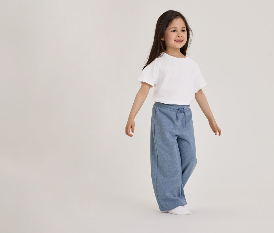 Ein junges Mädchen mit langen dunklen Haaren steht nach rechts blickend da und trägt ein weißes Kurzarm-T-Shirt, eine weite blaue Jogginghose mit Kordelzug und weiße Socken.