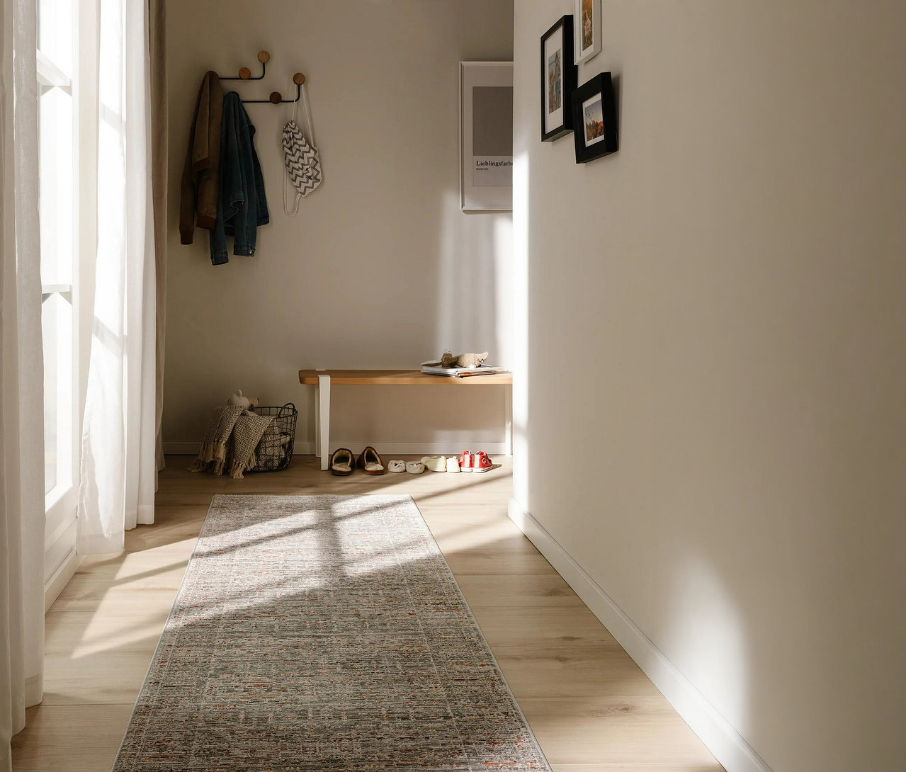 Heller Flur mit Holzboden und einem benuta Läufer »Anwar«, grau-orange. Sonnenlicht fällt durch ein Fenster auf den Läufer. An der Wand hängen Jacken an einer Garderobe und Bilder, darunter eine Holzbank mit Schuhen.