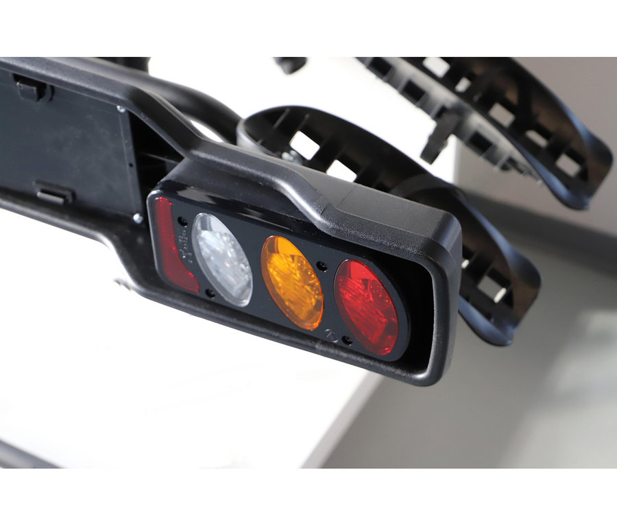Schwarzes Autozubehör, möglicherweise ein Fahrradträger oder eine Anhängerlichtleiste, mit Kennzeichenhalterung und einer Reihe von drei Lichtern: links rot, mittig klar/weiß und rechts bernsteinfarben/orange.