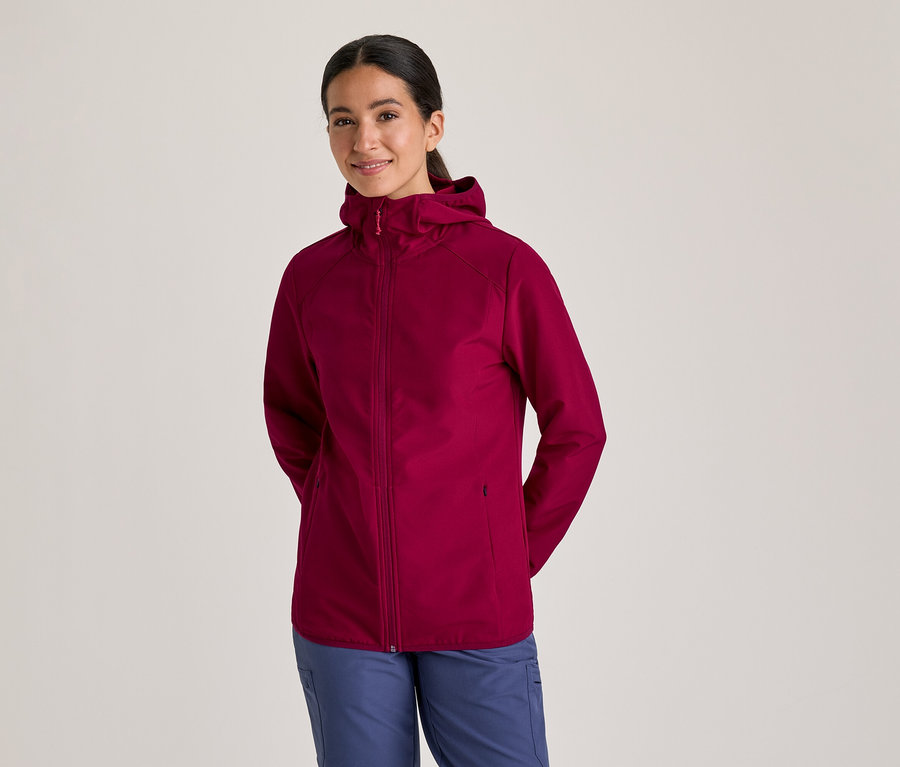 Frau in einer bordeauxroten Kapuzenjacke und blauen Hosen, die lächelt und die Hände hinter dem Rücken hält.