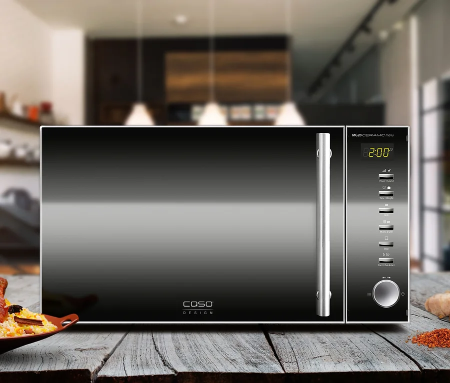 Schwarze Caso Design Mikrowelle mit silbernem Griff und digitaler Anzeige „2:00“ auf einem Holztisch. Links davon ein Teller mit Hähnchen und Reis, rechts Ingwer, Kurkuma, Zwiebel und Chiliflocken.